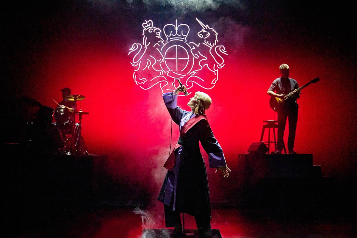 A singer in a robe performs dramatically under red lighting with a neon crest backdrop, while a guitarist and drummer play on raised platforms in the background.