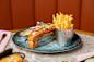 A gourmet lobster roll topped with herbs is served on a blue plate alongside a metal cup of golden fries and a side of sauce. The plate sits on a marble table, and a sign reads "burger & lobster.