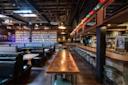 A rustic bar interior with dark wooden tables, green leather booths, a long wooden bar lined with stools, shelves of bottles, and string lights hanging from the ceiling. The space is empty and warmly lit.