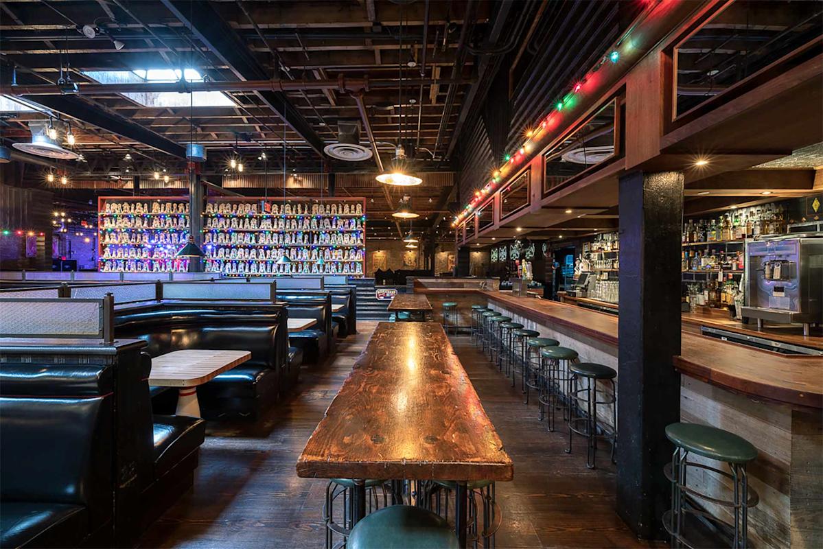 A rustic bar interior with dark wooden tables, green leather booths, a long wooden bar lined with stools, shelves of bottles, and string lights hanging from the ceiling. The space is empty and warmly lit.