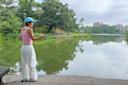 A person in a pink tank top and blue cap is fishing at a lakeside surrounded by trees. The sky is cloudy, and buildings are visible in the distance across the calm water.