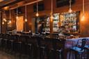 A stylish, empty bar with metal stools, pendant bulbs hanging above, and shelves stocked with various bottles of alcohol behind the wooden counter. The atmosphere is warm and inviting.
