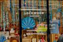 A colorful shop window display at Lockwood Sunnyside features hanging blue and turquoise decorations, a large shell cutout, gift items, books, and a mannequin wearing a light blue "QUEENS" t-shirt.