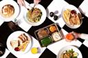 A variety of meals on a black and white checkered table. Dishes include salad, sandwiches with fries, chicken and waffles, a tray meal with veggies and mashed potatoes, and beverages like soda and juice. Hands are reaching for food and drinks.
