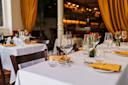 Elegant restaurant interior with tables set for dining, featuring white tablecloths, yellow napkins, glassware, small vases with red flowers, wooden chairs, and warm lighting with yellow curtains in the background.