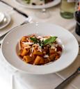 A white bowl of rigatoni pasta in tomato sauce, topped with grated cheese and fresh basil, sits on a white tablecloth next to a glass of red wine and a bottle of white wine.