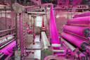 A textile factory interior shows machines processing bright pink fibers, with strands and dust covering equipment and the floor, illuminated by daylight from a window in the background.