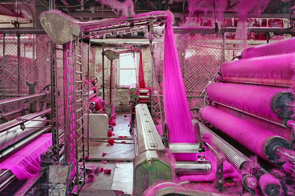 A textile factory interior shows machines processing bright pink fibers, with strands and dust covering equipment and the floor, illuminated by daylight from a window in the background.