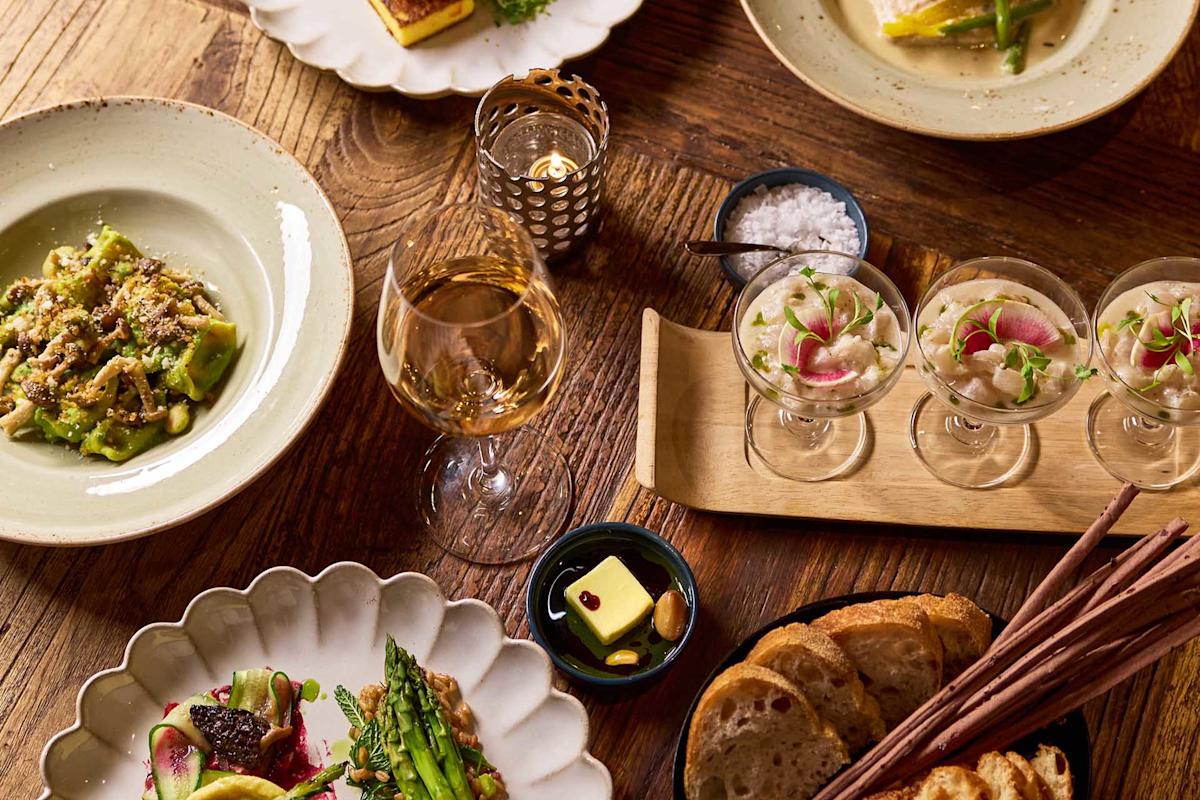 A wooden table set with assorted gourmet dishes, including pasta, vegetable plates, ceviche in glasses, a glass of white wine, bread slices, breadsticks, butter, and a bowl of salt, all lit by warm candlelight.