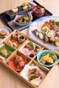 A colorful Japanese meal with assorted sushi, sashimi, tempura, grilled vegetables, and small dishes of seafood and vegetables, all beautifully arranged in a wooden bento box and on plates.