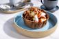 A waffle bowl filled with scoops of ice cream, caramelized pecans, and drizzled with caramel sauce, served on a beige and blue plate. A cup of coffee and small bowls of toppings are in the background on a marble table.