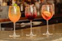 Three wine glasses filled with colorful drinks on a bar counter: an orange drink with lime, a dark red drink with ice, and a pink drink with a lemon slice, all garnished stylishly, set against a blurred bar background.