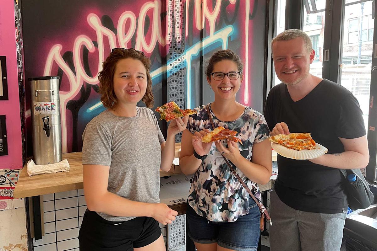 Three people smiling and holding slices of pizza inside a brightly decorated pizzeria with neon signs and large windows letting in natural light.