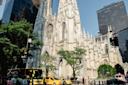 A vibrant city scene featuring St. Patrick's Cathedral in New York City. The gothic architecture stands tall amidst modern skyscrapers. Yellow taxis and pedestrians animate the street, and trees add greenery to the urban setting.