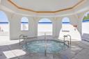 A circular hot tub with bubbling water is set in a bright, open pavilion with white walls, arched windows, and blue trim. Sunlight streams in, and stainless steel railings surround the tub.