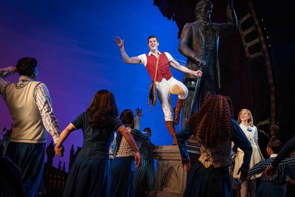 A man in a red vest and white pants poses energetically on a statue as a group of people in blue and white costumes reach toward him on stage, under bright theatrical lighting with a blue and purple background.