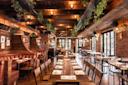 A cozy, warmly lit restaurant interior with brick walls and wooden beams. Tables are arranged with menus and place settings. Green plants hang from the ceiling, and sunlight streams in through large windows, creating a welcoming atmosphere.