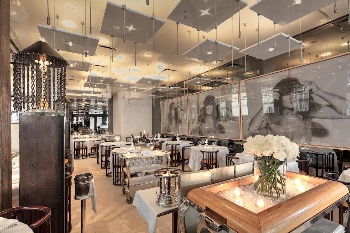 Elegant restaurant interior with neatly set tables, white tablecloths, modern lighting, large black-and-white portraits on the wall, and a centerpiece of white flowers and candles on a wooden cart in the foreground.