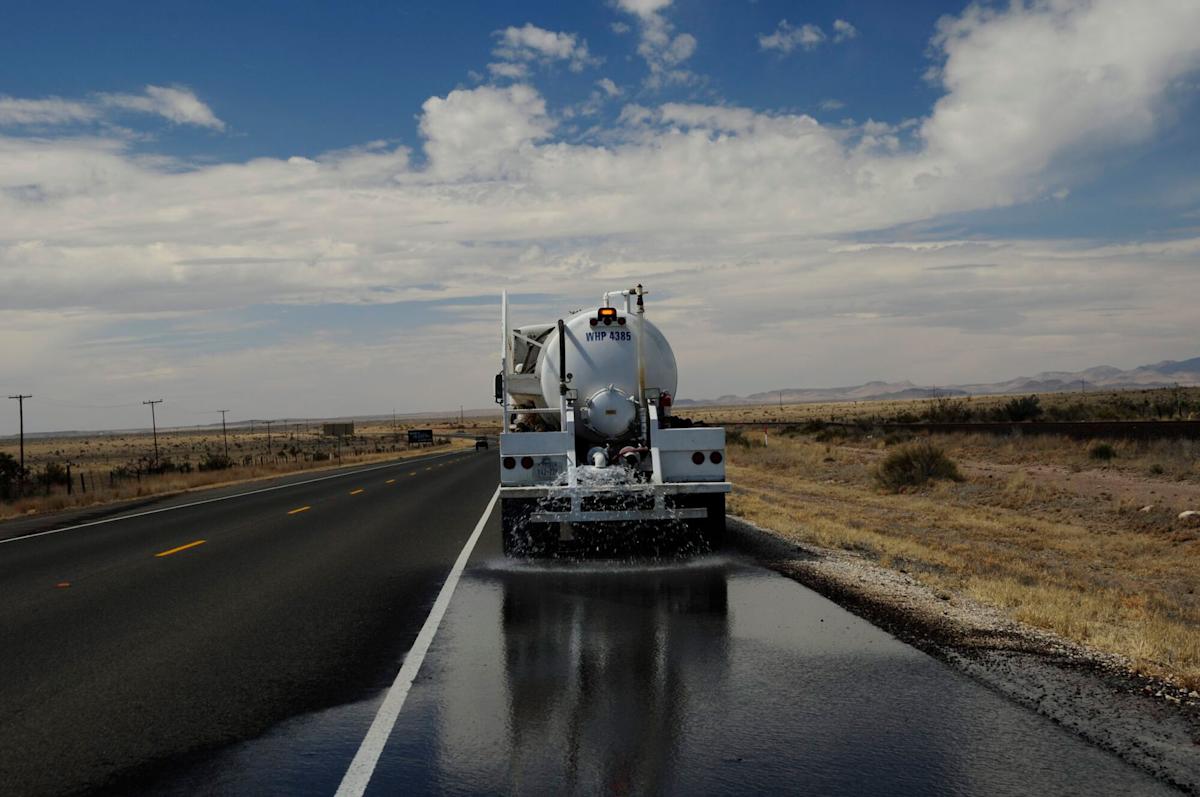 A water truck sprays water onto a road in a dry, desert landscape under a partly cloudy sky. The scene shows arid vegetation and distant mountains beside the highway.