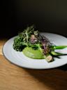 A white plate with a green vegetable dish, featuring broccolini, snap peas, leafy greens, sliced almonds, and a smooth green puree, set on a wooden table with a dark background.