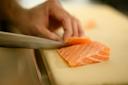A close-up of a hand slicing a piece of raw salmon with a sharp knife on a light-colored cutting board.