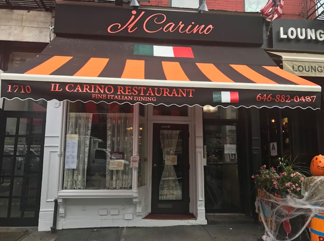 A storefront of Il Carino Restaurant with a brown and orange striped awning, Italian flag, and "Fine Italian Dining" written above the entrance. The door is flanked by windows and a small pumpkin sits outside.