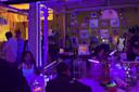 A vibrant room with neon and black lights shows people sitting at tables, art supplies scattered around. Colorful artwork and hearts cover the walls, while glowing decorations and creative activity give a lively, creative atmosphere.