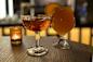 A close-up of two drinks on a wooden table: one in an ornate glass with a reddish-orange liquid, and a second in a rounded glass with an orange liquid garnished with a slice of citrus. A small lit candle is in the background.