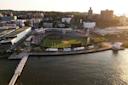 Aerial view of a riverside baseball stadium surrounded by urban buildings. The field is filled with spectators. A pier extends into the water, and the sun casts a warm glow over the scene.