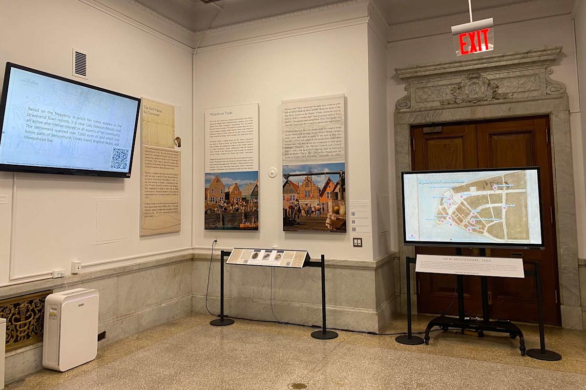 The image shows a museum exhibit featuring historical information and images about New Amsterdam. Two digital screens display text and maps on the walls, alongside large panels with text and images. An EXIT sign is visible above a door.