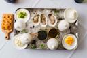 A tray with oysters on ice, surrounded by bowls containing caviar, sour cream, chopped eggs, green onions, and other garnishes. To the side, a small wooden board holds round toasted bread slices.