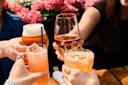 Four people clink glasses filled with various drinks—beer, wine, and cocktails—over a wooden table, with pink flowers in the background.
