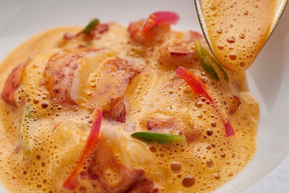 Close-up of a gourmet dish featuring pieces of seafood with thinly sliced red onions and green peppers, topped with a frothy, orange-hued sauce being spooned over the dish.