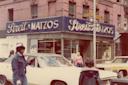 Street scene with people crossing near a vintage car in front of Streit’s Matzos, a bakery with a blue sign on a brick building. The historic corner location is marked by Suffolk and Rivington street signs. Pedestrians and urban ambiance are visible.