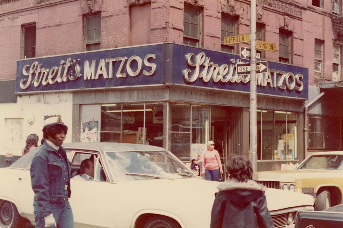 Street scene with people crossing near a vintage car in front of Streit’s Matzos, a bakery with a blue sign on a brick building. The historic corner location is marked by Suffolk and Rivington street signs. Pedestrians and urban ambiance are visible.