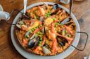 A round pan of paella with shrimp, mussels, clams, red pepper strips, and green peas on saffron rice. Three spoons are placed around the edges of the pan, which sits on a wooden table.