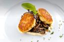 Two golden-brown crab cakes sit atop sautéed mushrooms, garnished with a leafy green and herbs on a white plate. A creamy sauce partially encircles the dish, adding a refined touch.