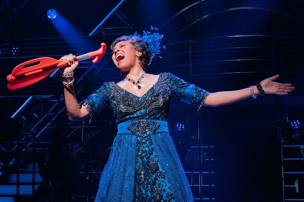 A performer in a blue dress and necklace passionately sings onstage, holding a large red prop shaped like a crutch, with dramatic lighting and stage equipment in the background.