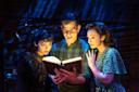 Three people in dramatic stage lighting look surprised as they read a glowing book. The person on the left wears dark, gothic clothing; the middle person wears glasses and plaid; the person on the right wears a green floral dress.