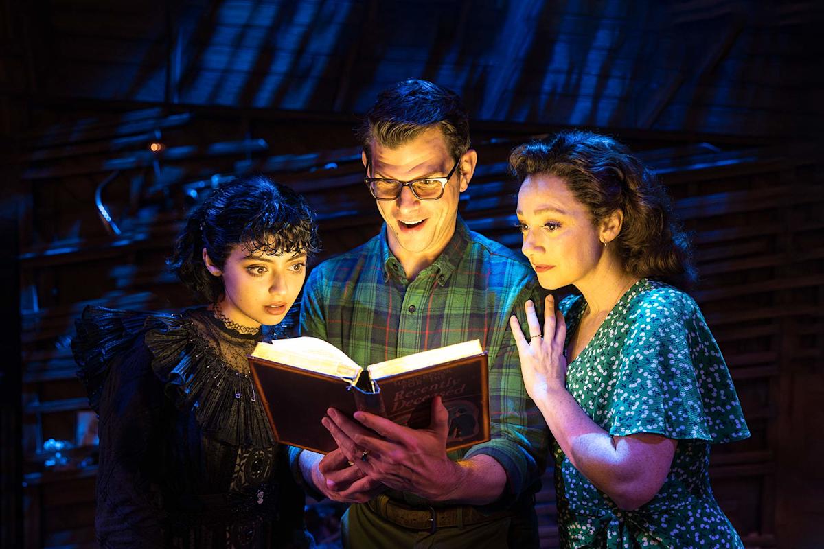 Three people in dramatic stage lighting look surprised as they read a glowing book. The person on the left wears dark, gothic clothing; the middle person wears glasses and plaid; the person on the right wears a green floral dress.