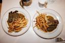 Two plates of steak with brown sauce and French fries on a white tablecloth, viewed from above. A glass of red wine, a small white pot, and part of a fork are also visible on the table.