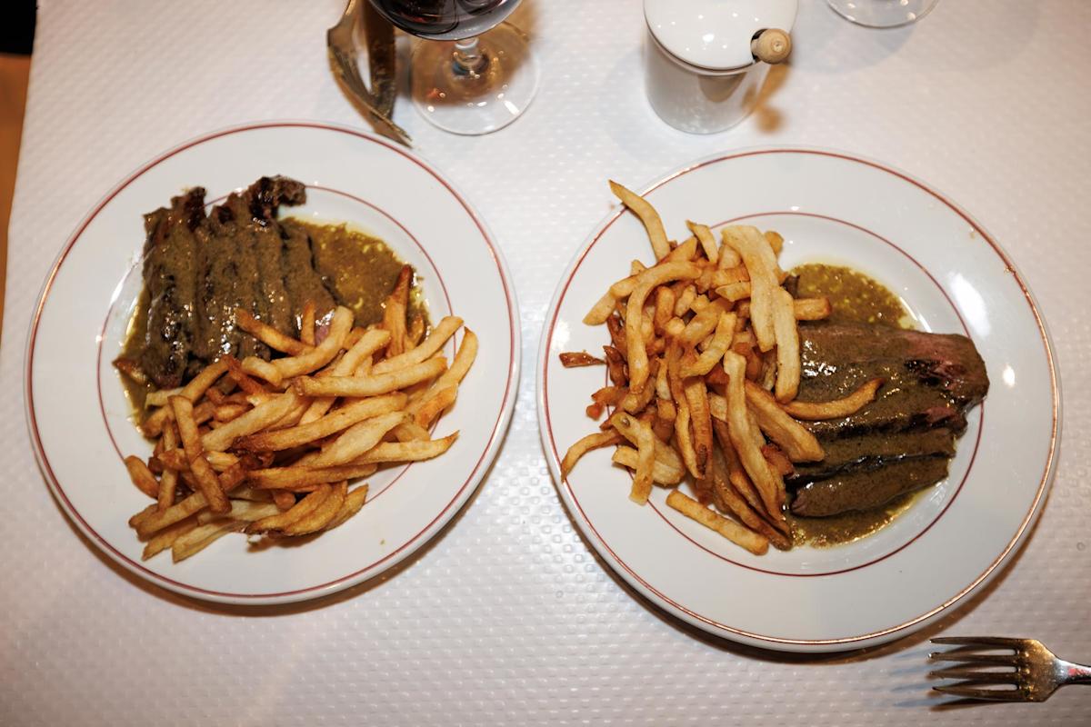 Two plates of steak with brown sauce and French fries on a white tablecloth, viewed from above. A glass of red wine, a small white pot, and part of a fork are also visible on the table.