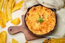 A skillet of baked cheese dip topped with chopped green onions sits on a wooden board, surrounded by yellow plantain chips and a light-colored cloth.