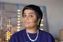 A person with short dark hair, wearing a purple top, a beaded necklace, and a green hoop earring, stands indoors with a blurred background of lights and signs, smiling gently at the camera.