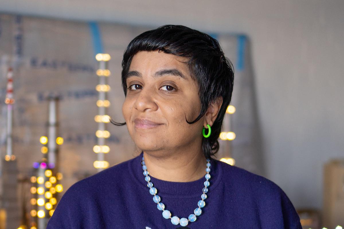 A person with short dark hair, wearing a purple top, a beaded necklace, and a green hoop earring, stands indoors with a blurred background of lights and signs, smiling gently at the camera.