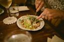 A person uses a knife and fork to cut into a salad topped with nuts or grains on a decorative plate. A glass of white wine and a glass of water are nearby on a wooden table.