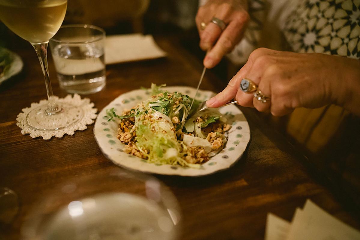 A person uses a knife and fork to cut into a salad topped with nuts or grains on a decorative plate. A glass of white wine and a glass of water are nearby on a wooden table.
