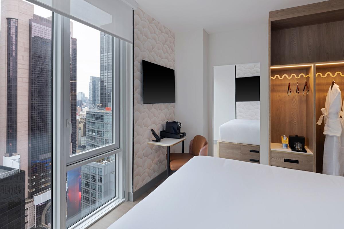 A modern hotel room with a large bed, wall-mounted TV, desk, open closet, and city view through floor-to-ceiling windows. A robe hangs in the closet, and tall buildings are visible outside.