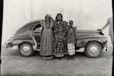 Three people in traditional patterned clothing stand in front of a vintage car. Two adults are in ornate dresses and headpieces, while a child stands beside them. The background is a bare wall, and part of another person is visible on the far right.