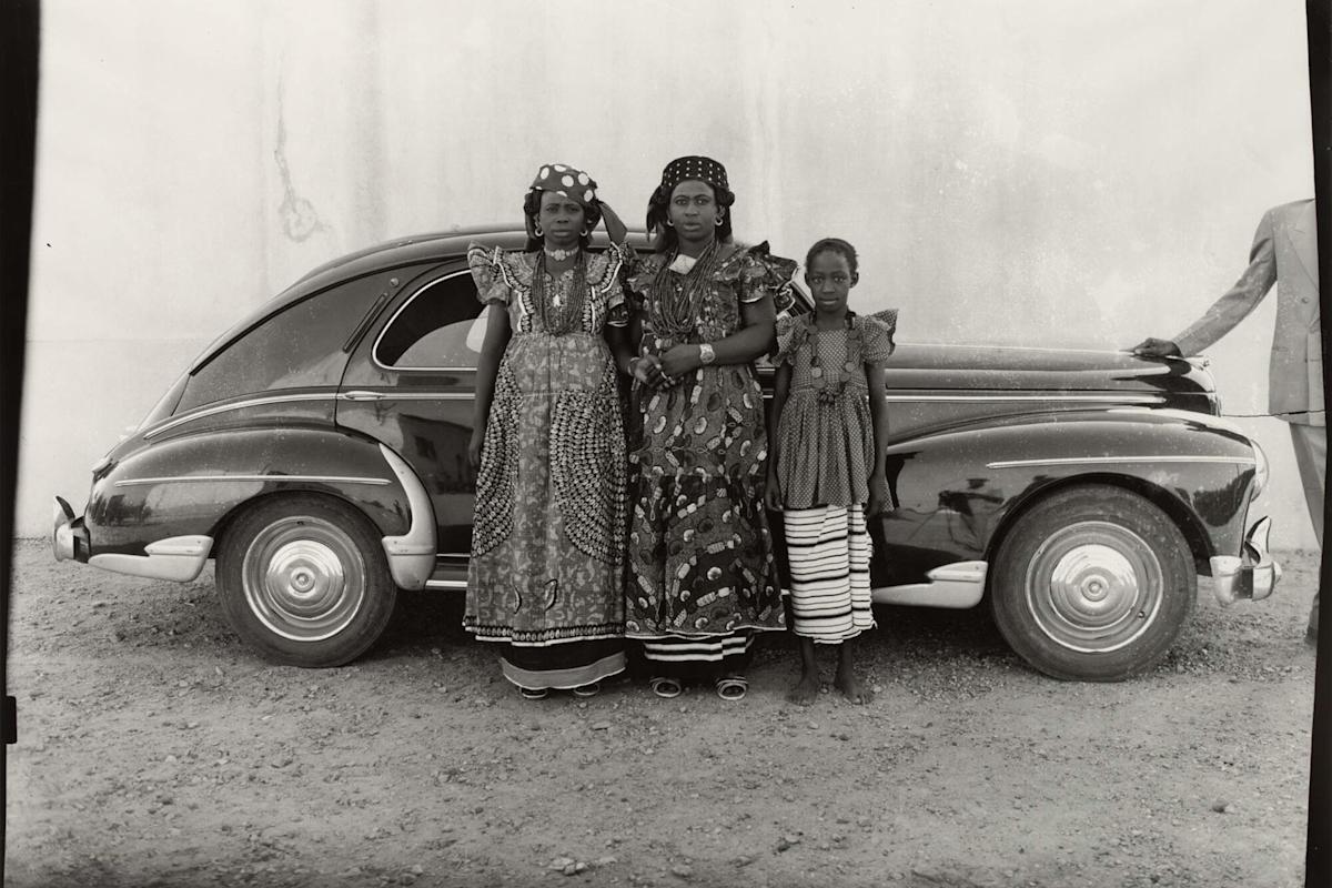 Three people in traditional patterned clothing stand in front of a vintage car. Two adults are in ornate dresses and headpieces, while a child stands beside them. The background is a bare wall, and part of another person is visible on the far right.